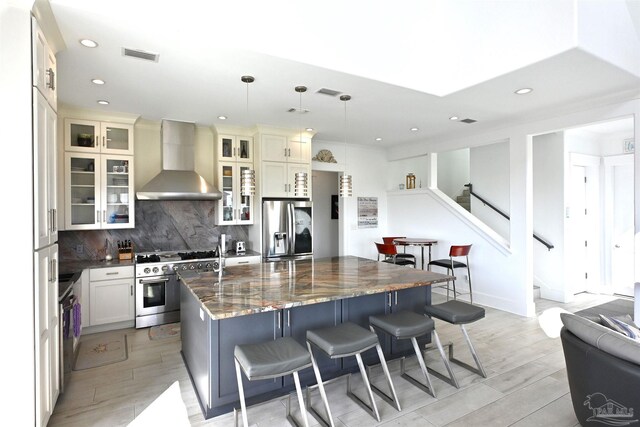 kitchen with stainless steel appliances, wall chimney range hood, light hardwood / wood-style floors, a kitchen island, and hanging light fixtures