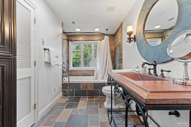 bathroom featuring stone tile floors, toilet, a sink, tiled shower / bath combo, and baseboards