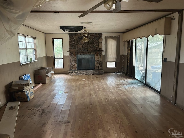 unfurnished living room featuring a textured ceiling, ceiling fan, hardwood / wood-style floors, a stone fireplace, and wood walls