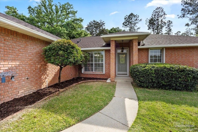 entrance to property featuring brick siding, a yard, and roof with shingles