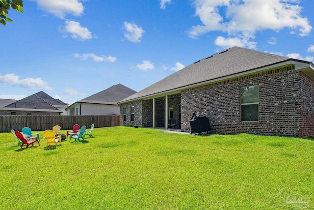 rear view of property featuring fence, roof with shingles, brick siding, an outdoor fire pit, and a yard