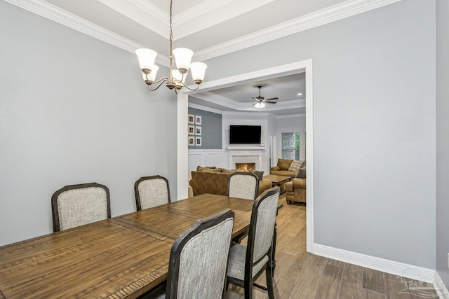 dining space with a raised ceiling, ceiling fan with notable chandelier, baseboards, a lit fireplace, and ornamental molding