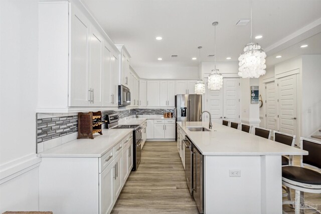 kitchen featuring a kitchen island with sink, hanging light fixtures, stainless steel appliances, sink, and white cabinetry