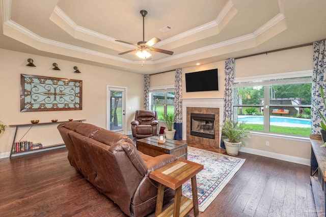 living room featuring a tiled fireplace, crown molding, a raised ceiling, dark hardwood / wood-style flooring, and ceiling fan