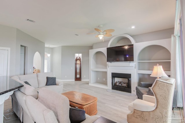 living room with a tile fireplace, light wood-type flooring, built in shelves, and ceiling fan