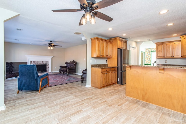 kitchen with ceiling fan, light hardwood / wood-style flooring, dark stone countertops, a fireplace, and stainless steel refrigerator