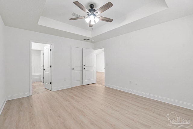 unfurnished bedroom featuring a raised ceiling, ensuite bathroom, ceiling fan, and light hardwood / wood-style floors