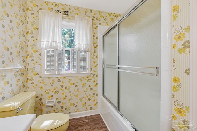 bathroom with wood finished floors, baseboards, shower / bath combination with glass door, wallpapered walls, and toilet