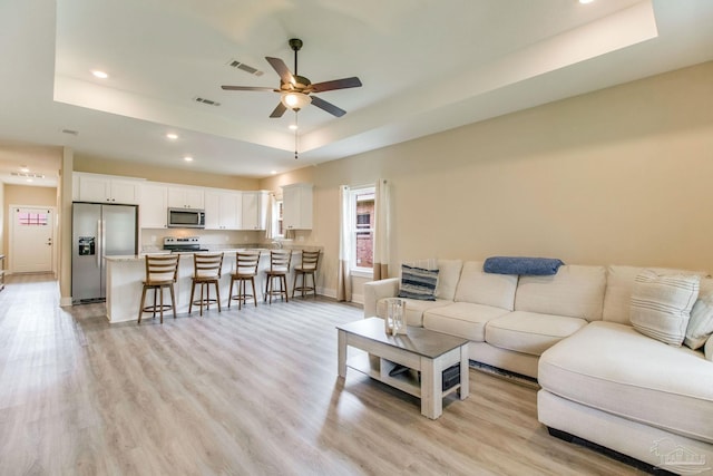 living room featuring a raised ceiling, ceiling fan, recessed lighting, light wood finished floors, and baseboards