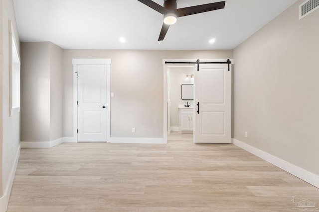 unfurnished bedroom featuring light wood-type flooring, a barn door, ensuite bath, and ceiling fan