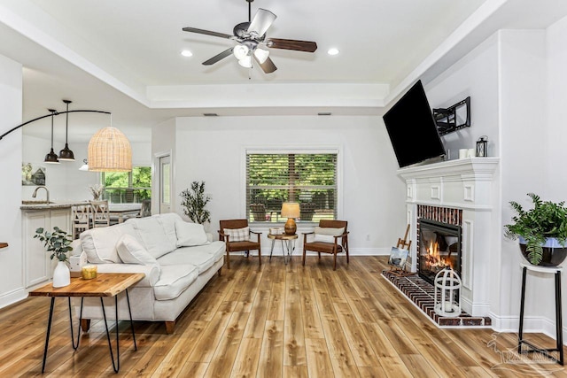 living area with recessed lighting, a tray ceiling, a ceiling fan, light wood-type flooring, and a brick fireplace