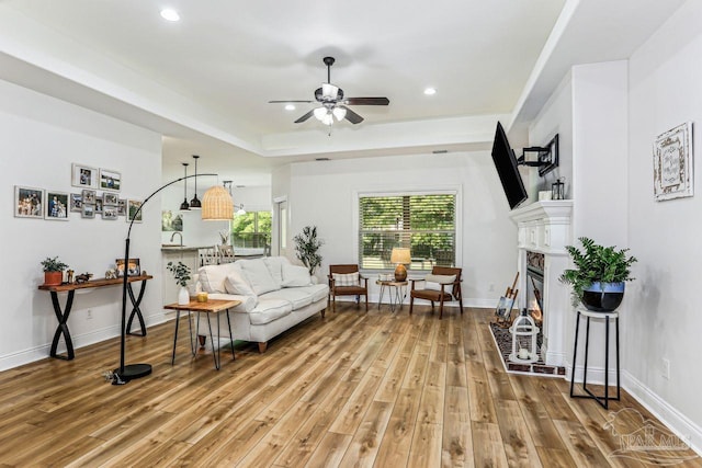 living area featuring light wood-style flooring, a raised ceiling, recessed lighting, ceiling fan, and a lit fireplace
