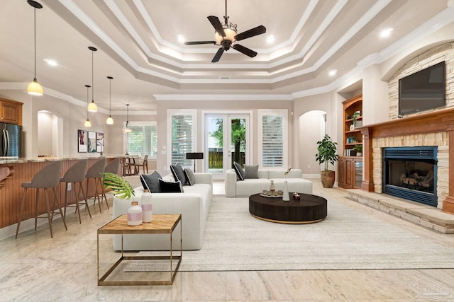 living area with built in features, a fireplace with raised hearth, ceiling fan, ornamental molding, and a tray ceiling