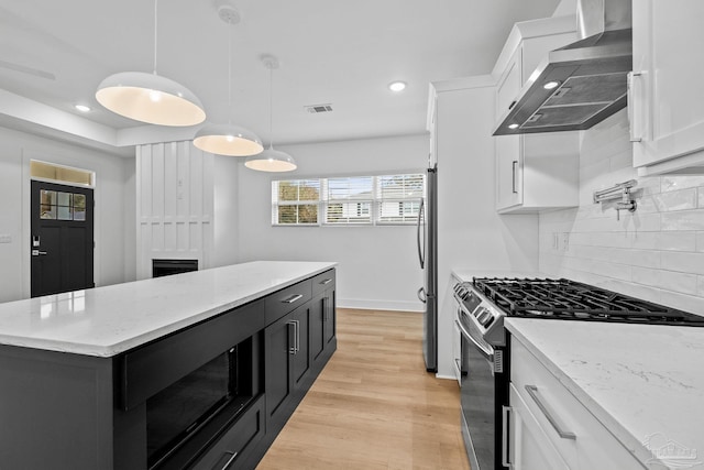 kitchen featuring wall chimney range hood, appliances with stainless steel finishes, white cabinetry, a center island, and decorative light fixtures