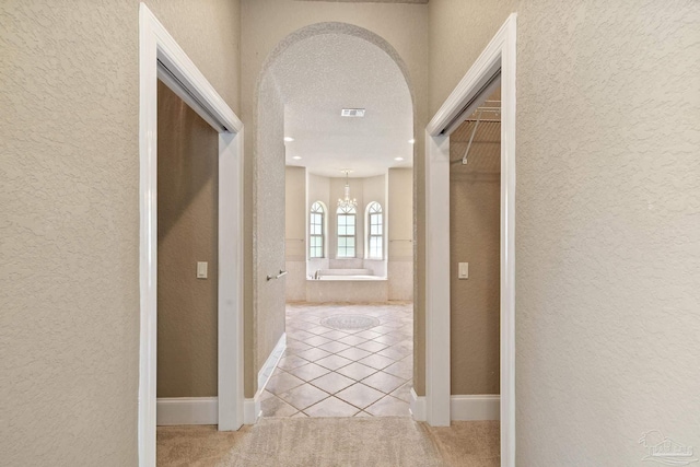 hallway featuring a textured wall, light tile patterned flooring, a chandelier, light carpet, and arched walkways