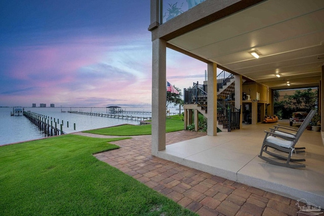 patio terrace at dusk with a water view, a dock, and a lawn