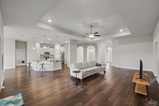 living area with baseboards, a ceiling fan, dark wood finished floors, arched walkways, and a tray ceiling