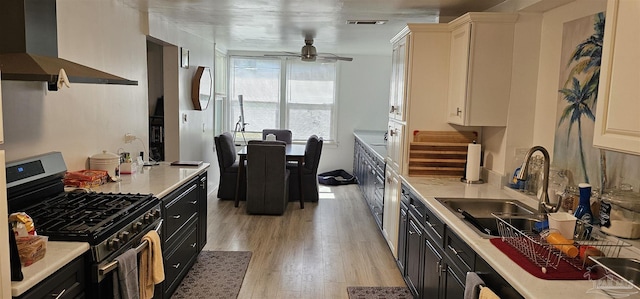 kitchen featuring wall chimney range hood, sink, ceiling fan, stainless steel gas stove, and light wood-type flooring