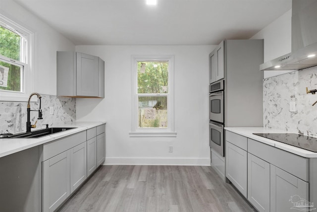 kitchen featuring gray cabinetry, backsplash, wall chimney range hood, sink, and light wood-type flooring