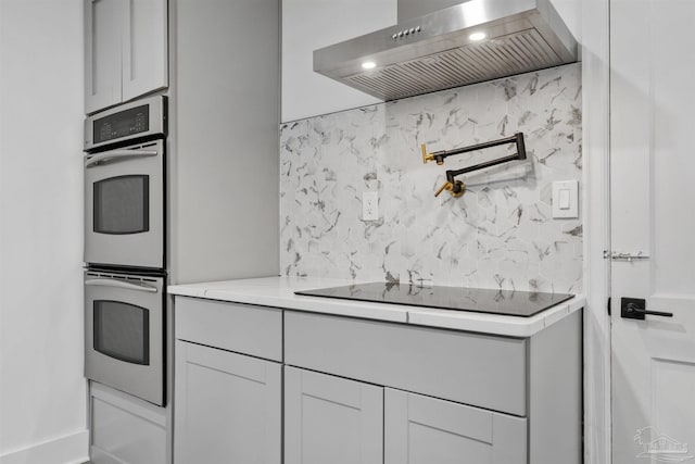 kitchen with light stone countertops, black electric stovetop, backsplash, double oven, and wall chimney range hood