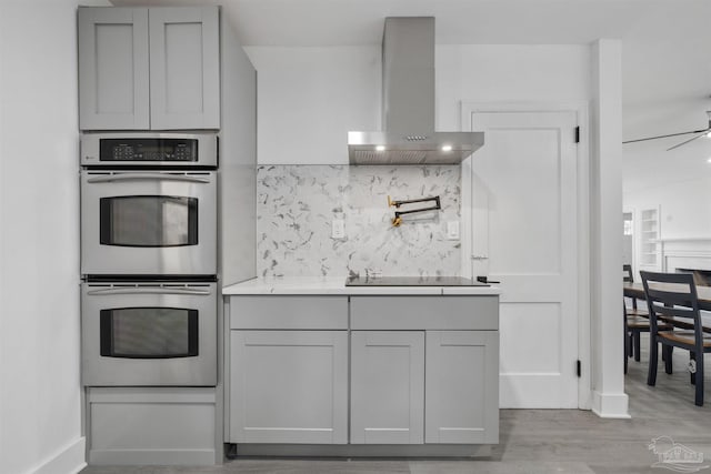 kitchen featuring black electric stovetop, wall chimney range hood, gray cabinets, light wood-type flooring, and double oven