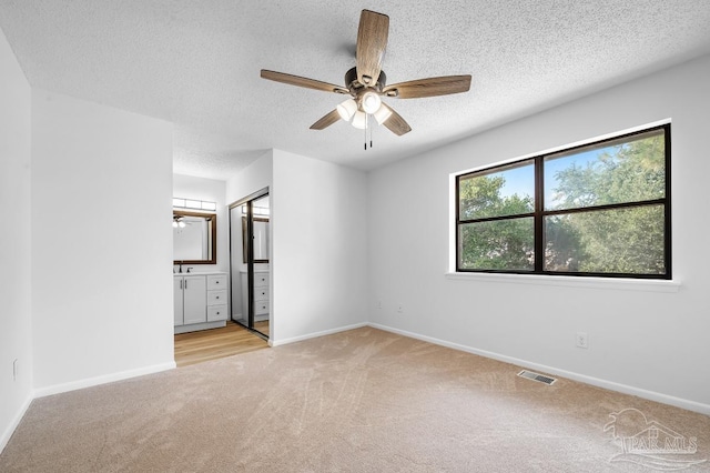 unfurnished bedroom with a textured ceiling, ensuite bath, light carpet, a closet, and ceiling fan