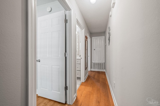 corridor featuring a textured wall, a textured ceiling, visible vents, light wood finished floors, and baseboards
