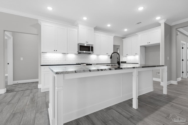 kitchen with backsplash, a kitchen island with sink, ornamental molding, light hardwood / wood-style floors, and white cabinetry