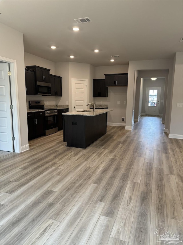 kitchen featuring sink, stainless steel appliances, light stone counters, an island with sink, and light wood-type flooring