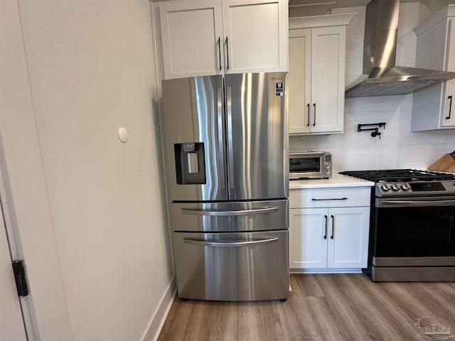 kitchen with appliances with stainless steel finishes, white cabinetry, wall chimney range hood, backsplash, and light hardwood / wood-style flooring