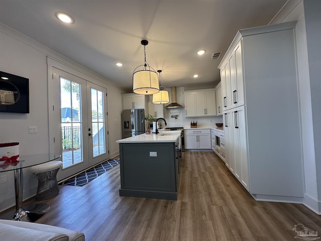 kitchen featuring pendant lighting, stainless steel refrigerator with ice dispenser, white cabinetry, wall chimney range hood, and a kitchen island with sink