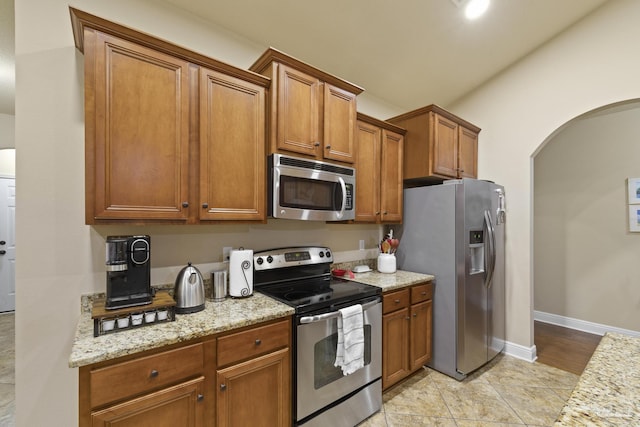 kitchen featuring baseboards, brown cabinetry, arched walkways, light stone counters, and stainless steel appliances