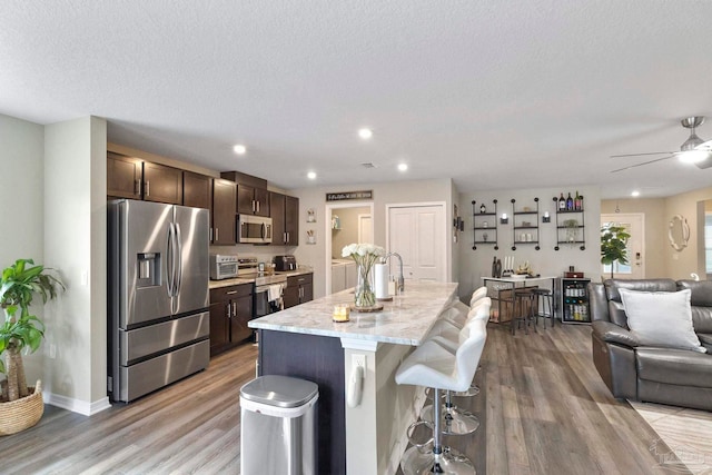 kitchen featuring stainless steel appliances, dark brown cabinetry, an island with sink, wood-type flooring, and ceiling fan