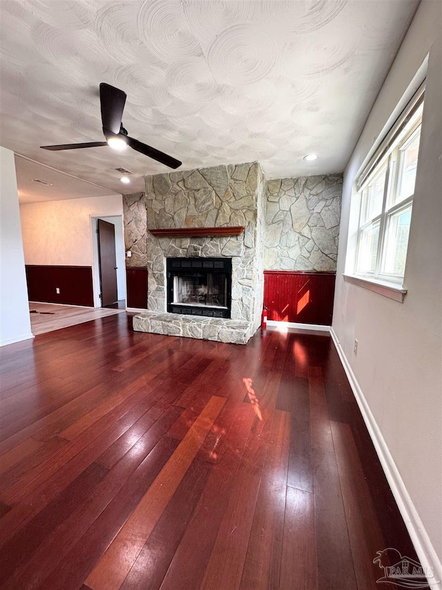 unfurnished living room with a textured ceiling, hardwood / wood-style floors, a stone fireplace, baseboards, and ceiling fan