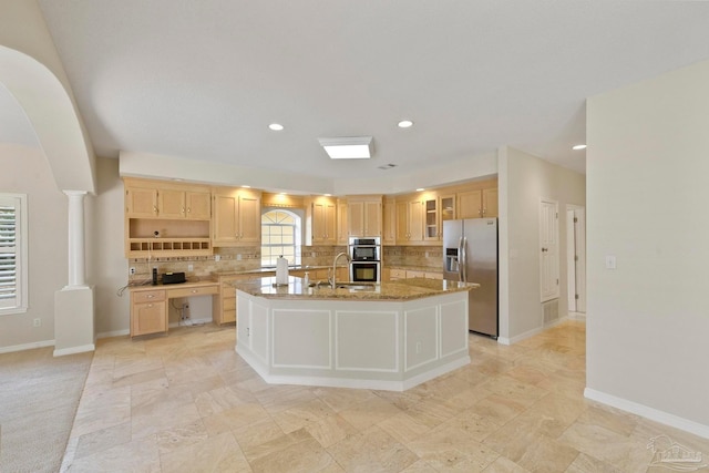 kitchen featuring a kitchen island with sink, stainless steel appliances, light stone countertops, light brown cabinetry, and ornate columns