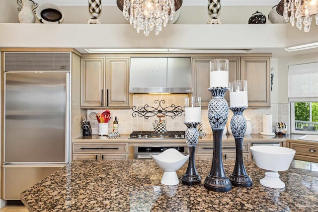 kitchen featuring a notable chandelier, dark stone counters, stainless steel appliances, wall chimney exhaust hood, and decorative backsplash