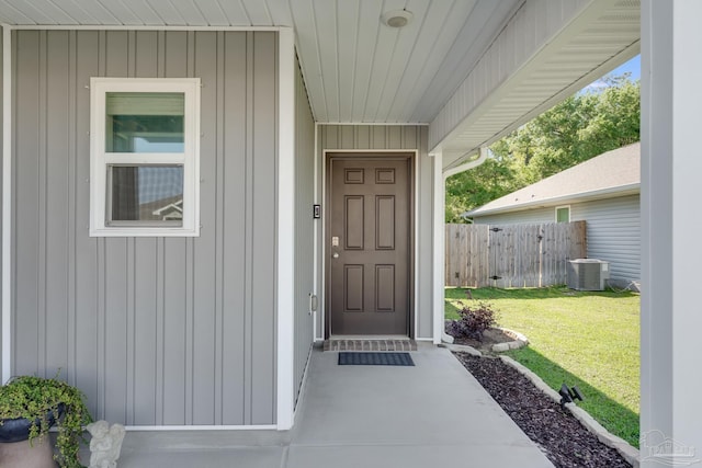 view of exterior entry with board and batten siding, a yard, central AC, and fence