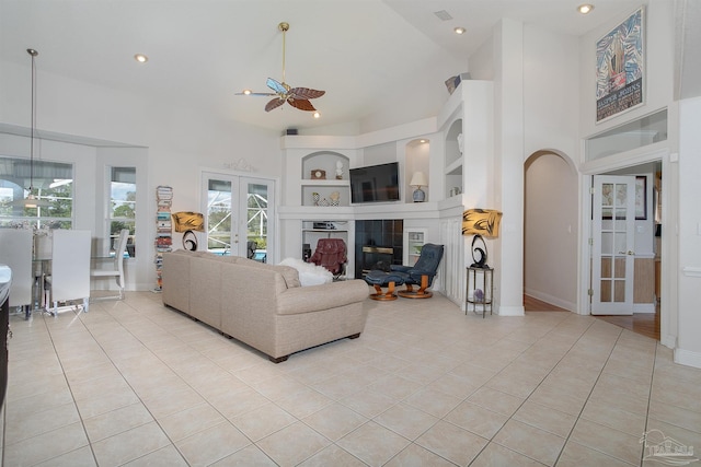 tiled living room featuring a tiled fireplace, built in shelves, ceiling fan, high vaulted ceiling, and french doors