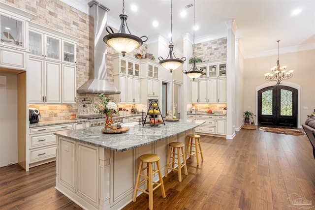 kitchen with decorative light fixtures, wall chimney range hood, decorative backsplash, a center island with sink, and dark wood-type flooring