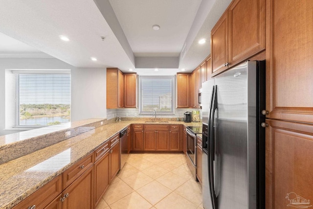 kitchen featuring brown cabinetry, light stone countertops, a tray ceiling, stainless steel appliances, and a sink