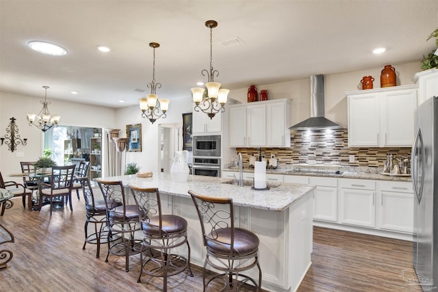 kitchen with white cabinetry, a kitchen island with sink, stainless steel appliances, decorative light fixtures, and wall chimney range hood