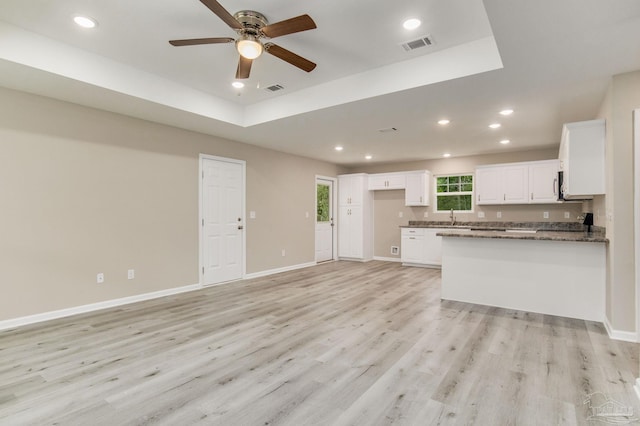 kitchen with light wood-type flooring, dark stone counters, a tray ceiling, ceiling fan, and white cabinetry