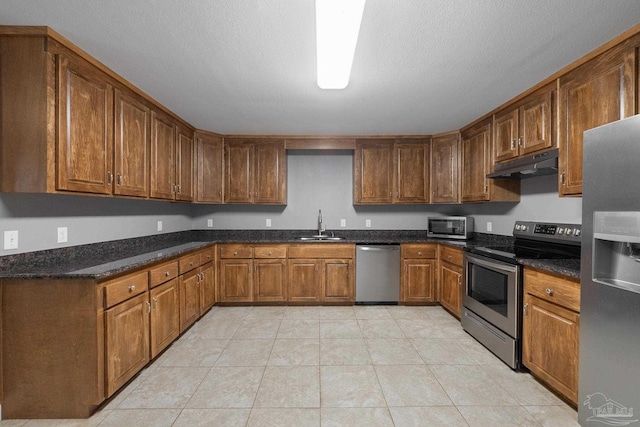 kitchen with brown cabinetry, under cabinet range hood, a sink, light tile patterned floors, and stainless steel appliances
