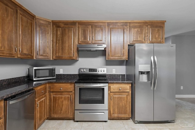 kitchen with brown cabinetry, appliances with stainless steel finishes, light tile patterned floors, dark stone counters, and under cabinet range hood