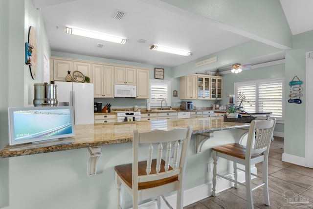 kitchen with light stone countertops, white appliances, kitchen peninsula, cream cabinets, and a breakfast bar