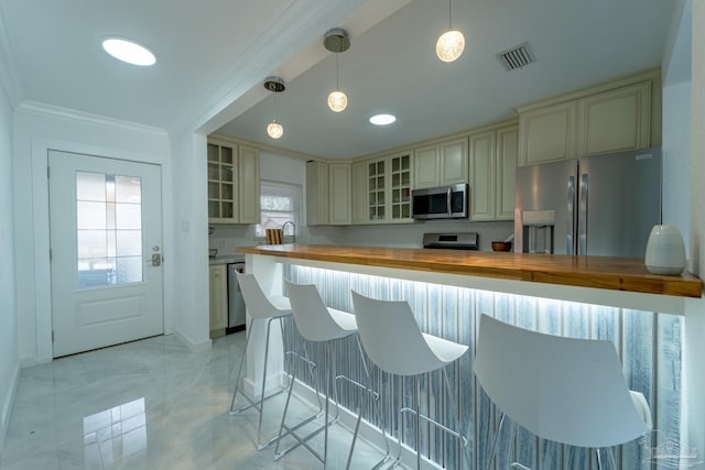 kitchen featuring crown molding, butcher block counters, stainless steel appliances, decorative light fixtures, and cream cabinetry