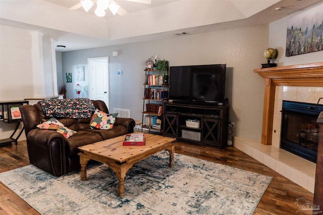 living room featuring a tile fireplace, ceiling fan, decorative columns, a tray ceiling, and dark hardwood / wood-style flooring