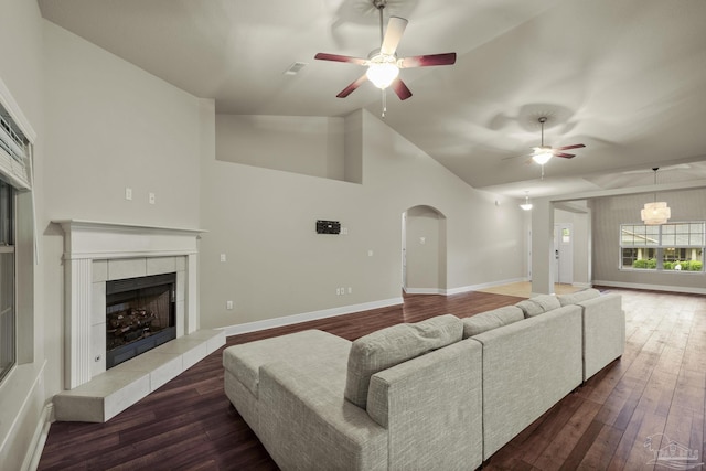 living area featuring ceiling fan, dark wood-type flooring, a tiled fireplace, arched walkways, and high vaulted ceiling