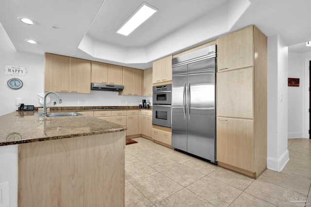 kitchen with a peninsula, stainless steel appliances, light brown cabinetry, under cabinet range hood, and a sink