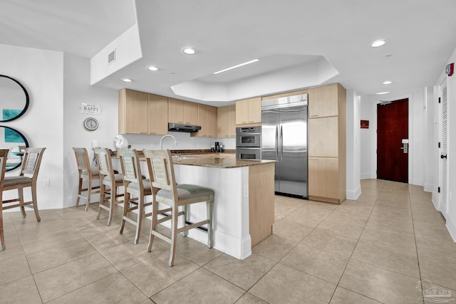 kitchen with light brown cabinets, a peninsula, stainless steel appliances, under cabinet range hood, and a kitchen bar